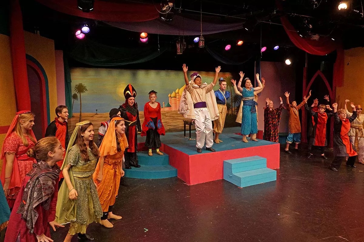 A theatrical performance with actors in colorful costumes on stage, hands raised, under vibrant lighting.