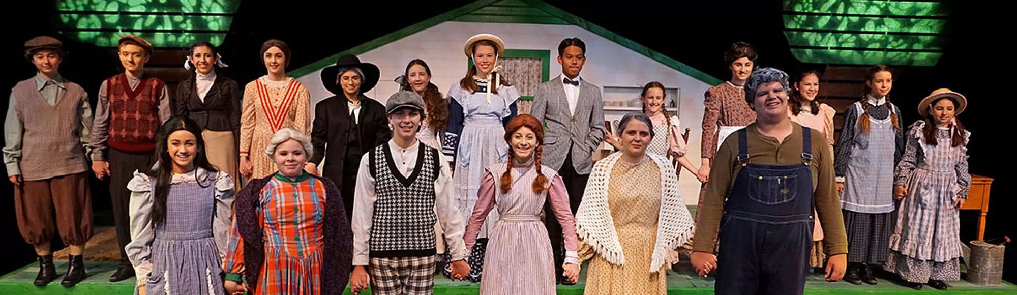 A group of people in historical costumes are holding hands on a stage set with a house backdrop, likely a theatrical performance.