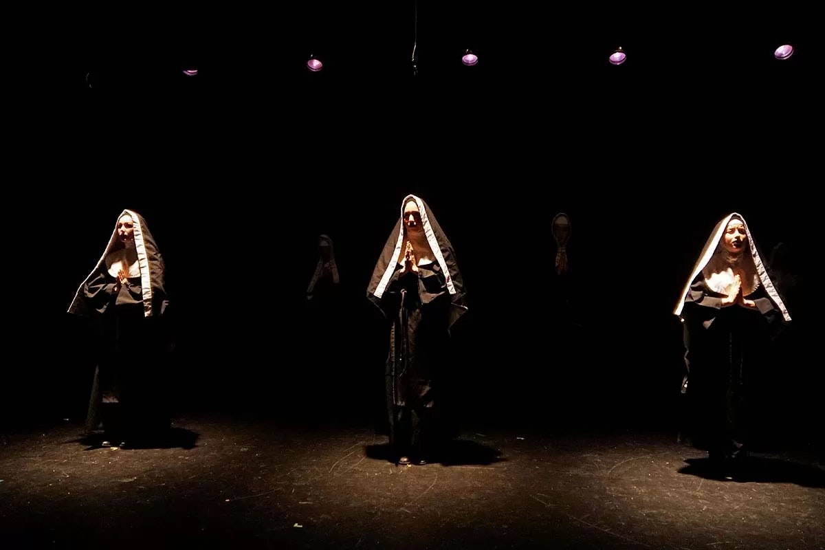 Three people dressed as nuns are standing under spotlights, in a dark setting.