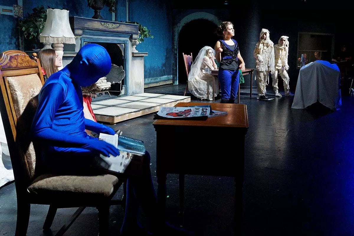 A theatrical scene with a person in a blue suit reading at a desk, others are dressed in various costumes on stage.