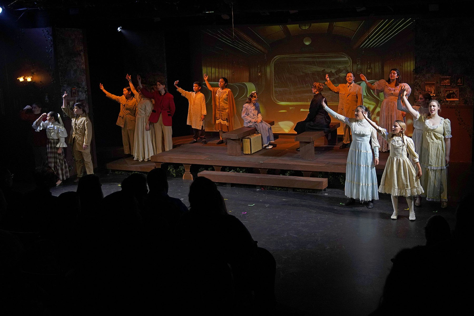 People in period costumes perform on stage with set resembling a train interior, under stage lights.