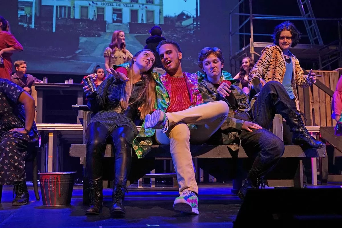 A group of people in colorful outfits are performing on stage, sitting and posing expressively with a projection in the background.