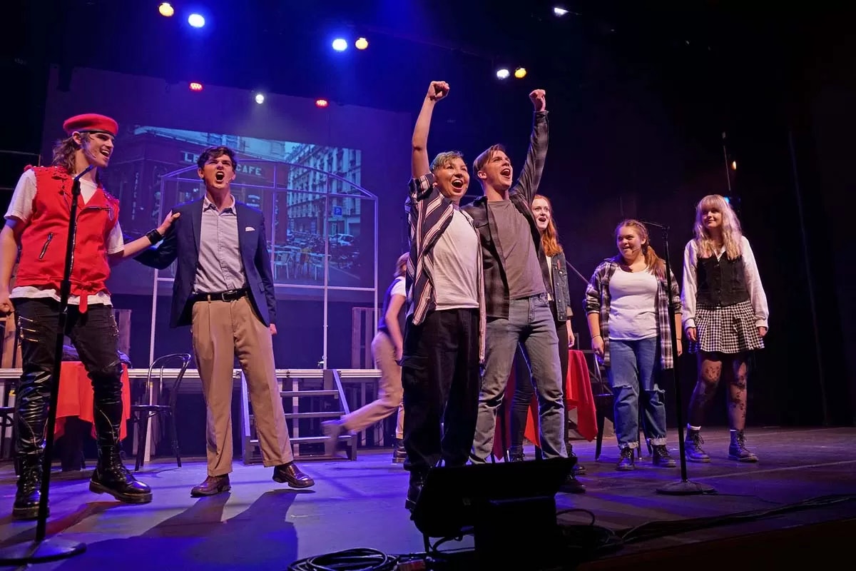 A group of people performing a musical on stage, with colorful lighting and a backdrop of a café scene.