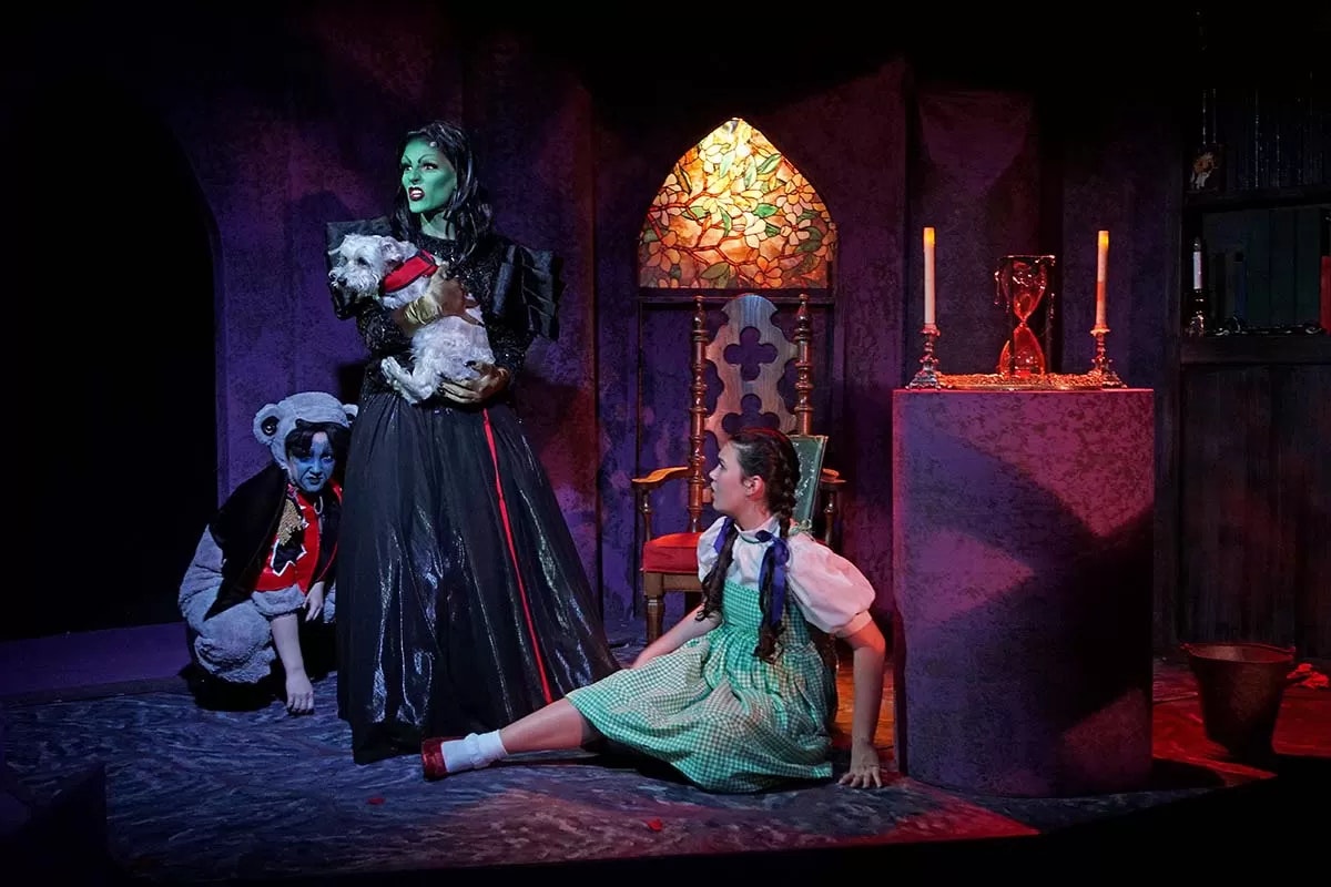 A theatrical scene with a green witch holding a dog, a girl in a green dress on the floor, and a figure with a blue face nearby.