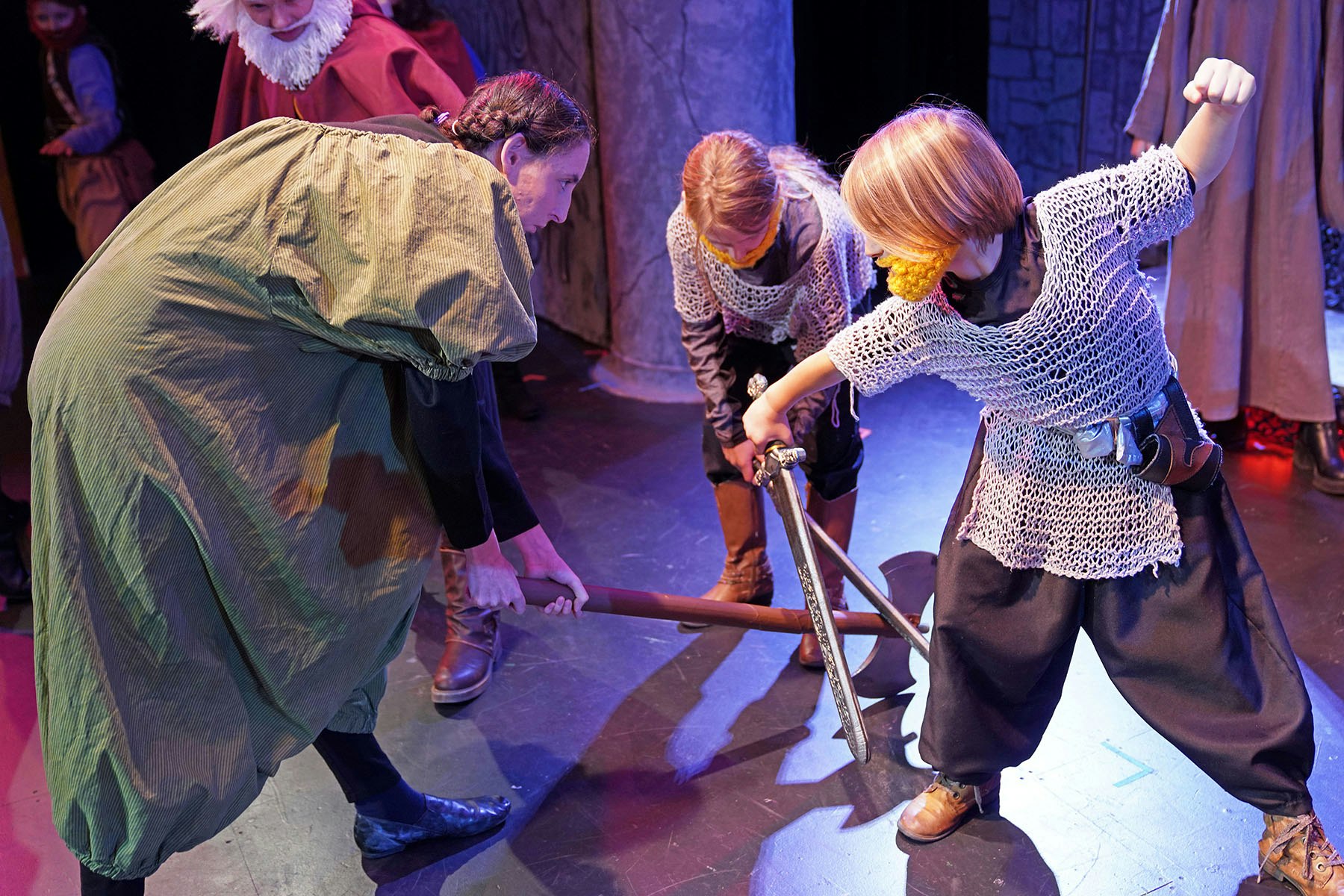 People in medieval costumes engaged in a staged sword and axe battle on a theater stage.