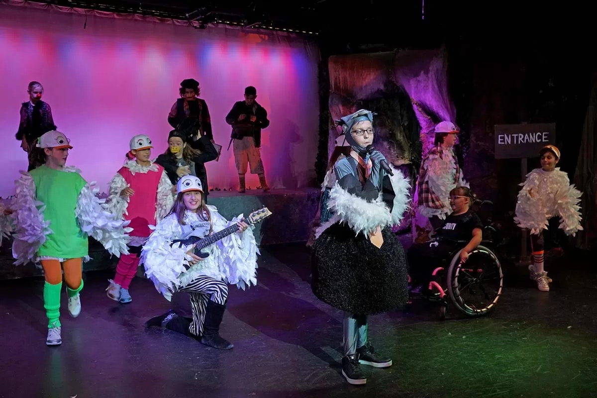 A group of children in colorful costumes perform on stage, some in bird outfits, one playing a guitar, and another in a wheelchair.