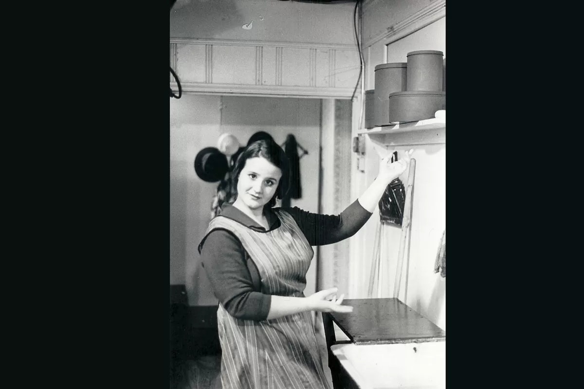 A woman in a striped apron is standing in a kitchen, lifting a string from a piece of furniture, with hats hanging in the background.