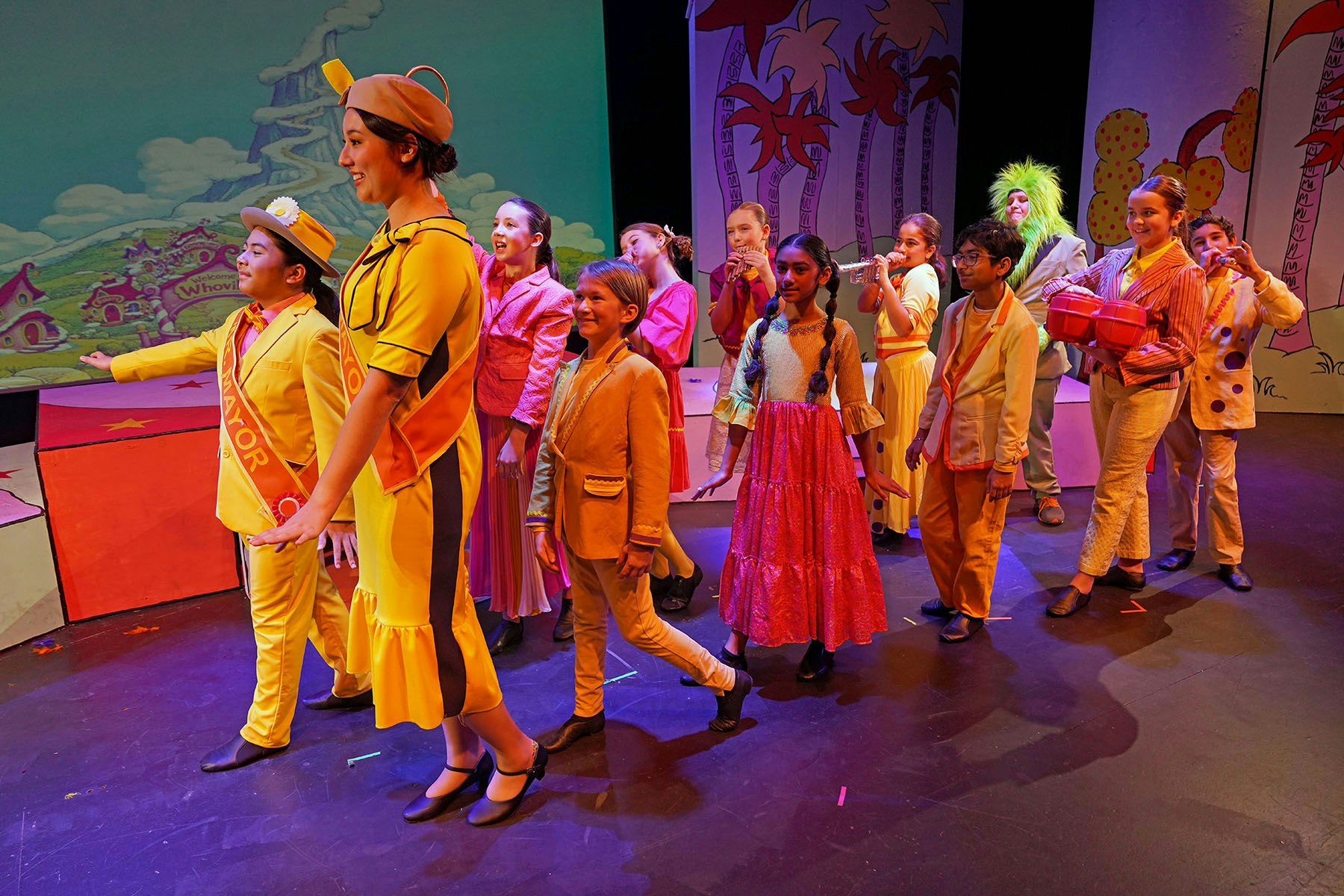 A colorful stage performance featuring children in vibrant costumes, with a whimsical backdrop, likely from a musical or play.