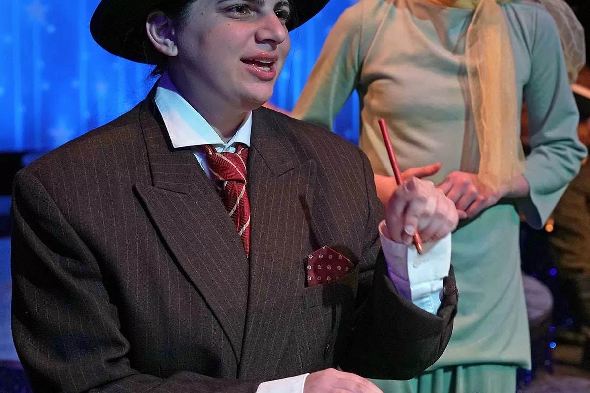 A person in a pinstripe suit and hat, holding a pencil, with another person in green behind them.