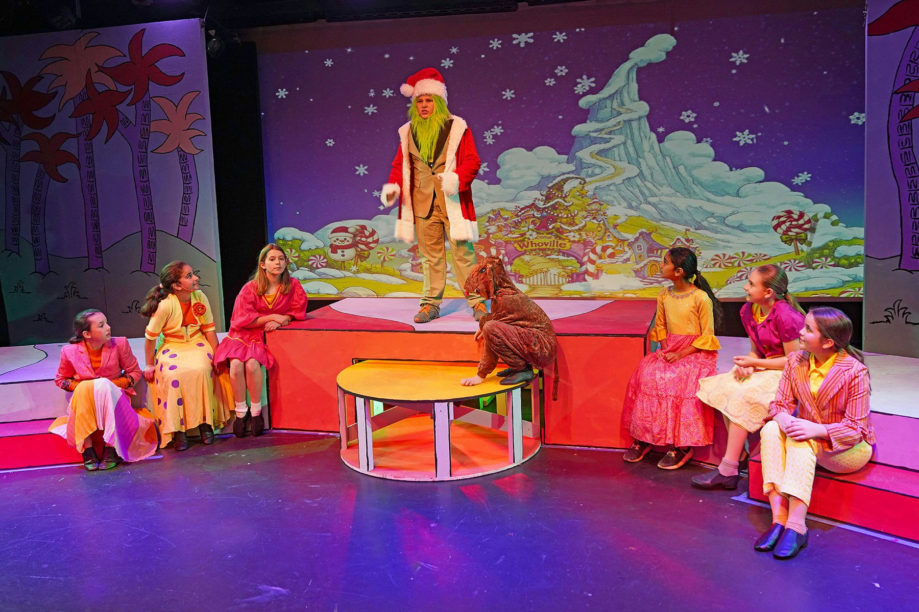 A festive stage scene featuring a character in a Santa outfit, children in colorful costumes, and a whimsical backdrop.
