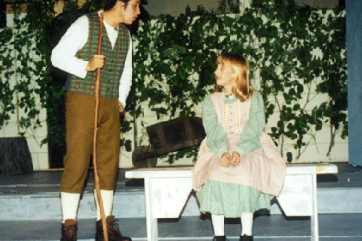 Two people in old-fashioned costumes; one holding a stick, the other sitting on a bench, in front of ivy-covered walls.
