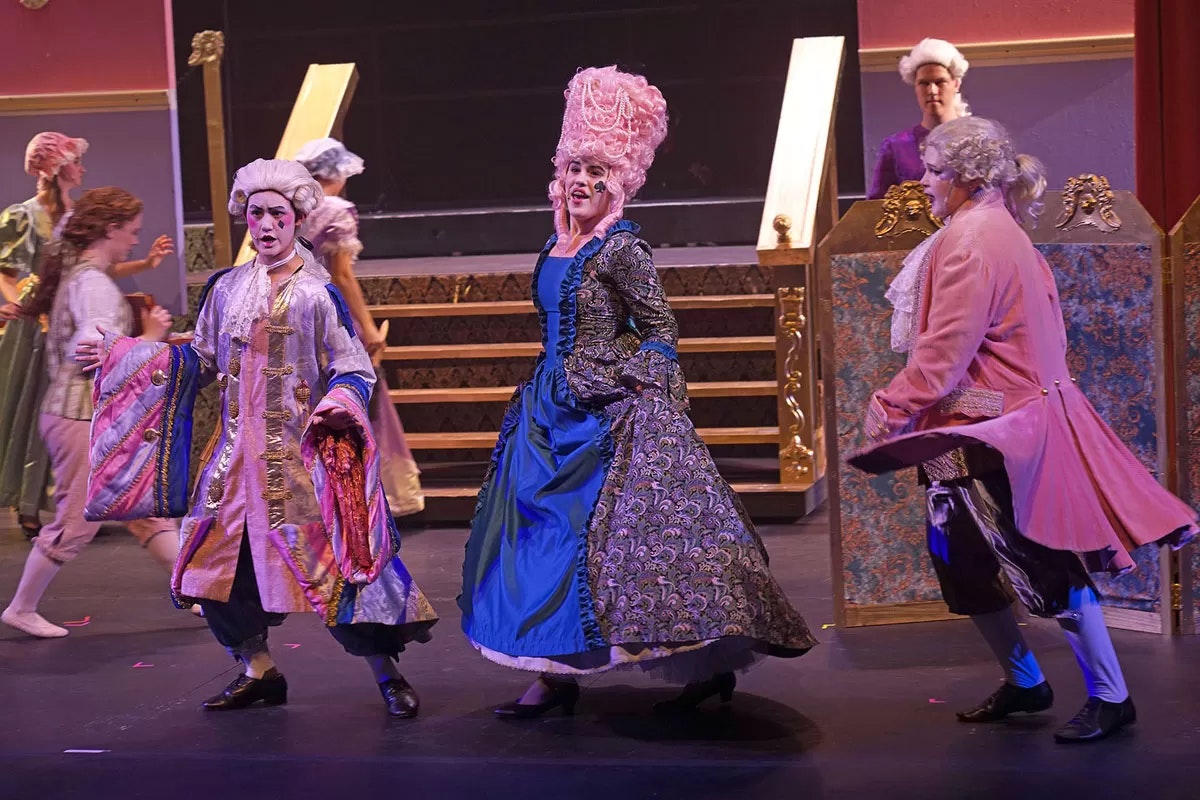 People in elaborate, historical costumes performing on stage with colorful attire and powdered wigs.