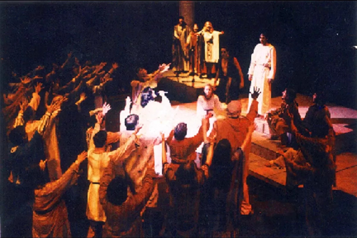 A theatrical performance with actors in robes, some raising hands, others watching from a higher platform.