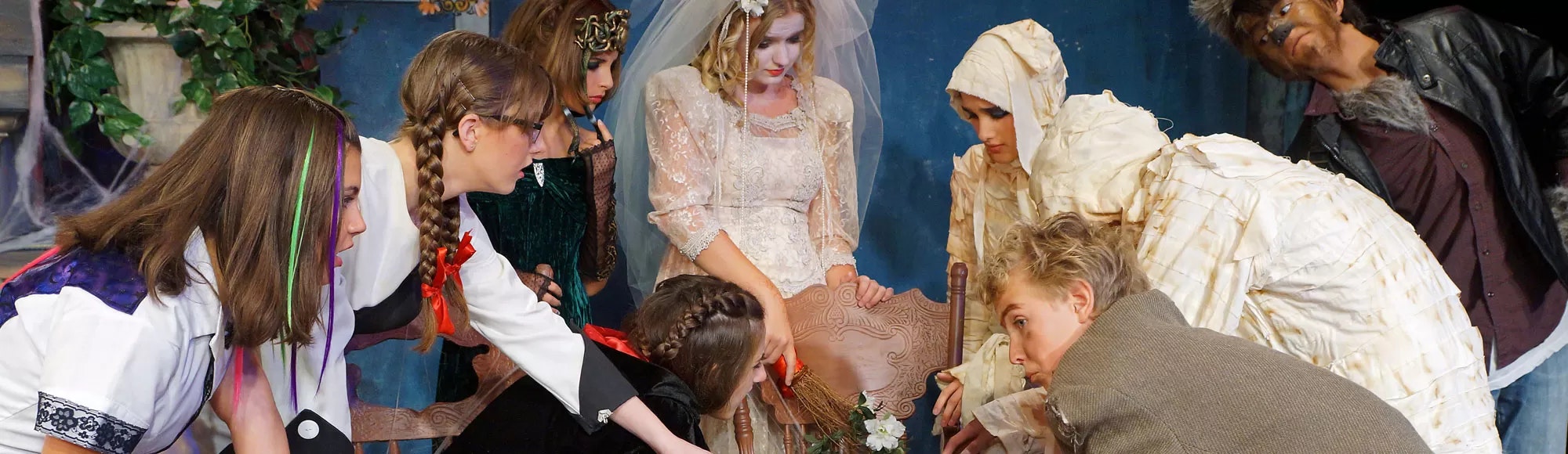 A group of people in costume gather around a chair holding a broom, revealing a theatrical or themed gathering.