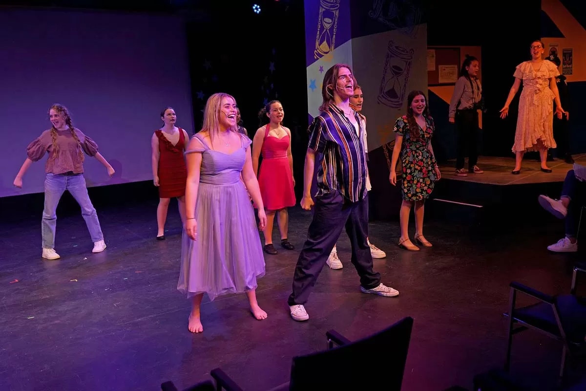 A group of performers on stage, energetically singing or acting, with colorful costumes and a backdrop.