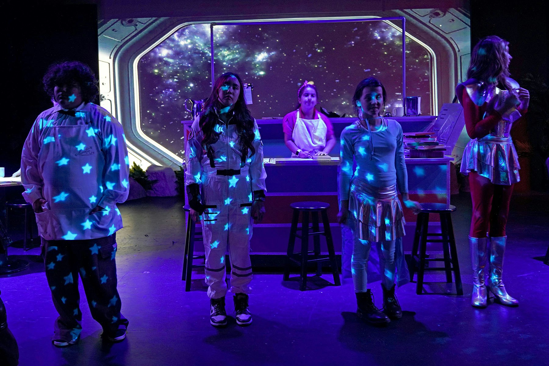 Five people in a space-themed stage setting with projected stars, wearing costumes including astronaut suits and futuristic outfits.