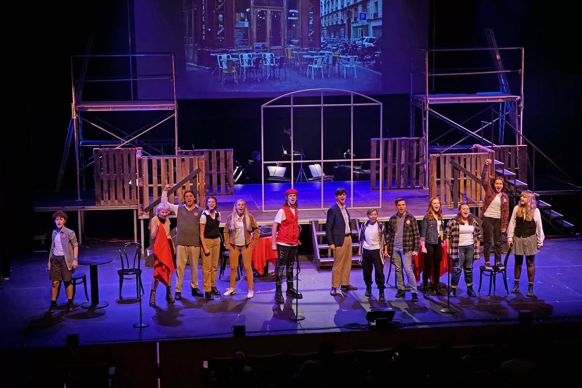 A group of performers on stage, singing, with a scaffold backdrop and a cafe scene projected above.