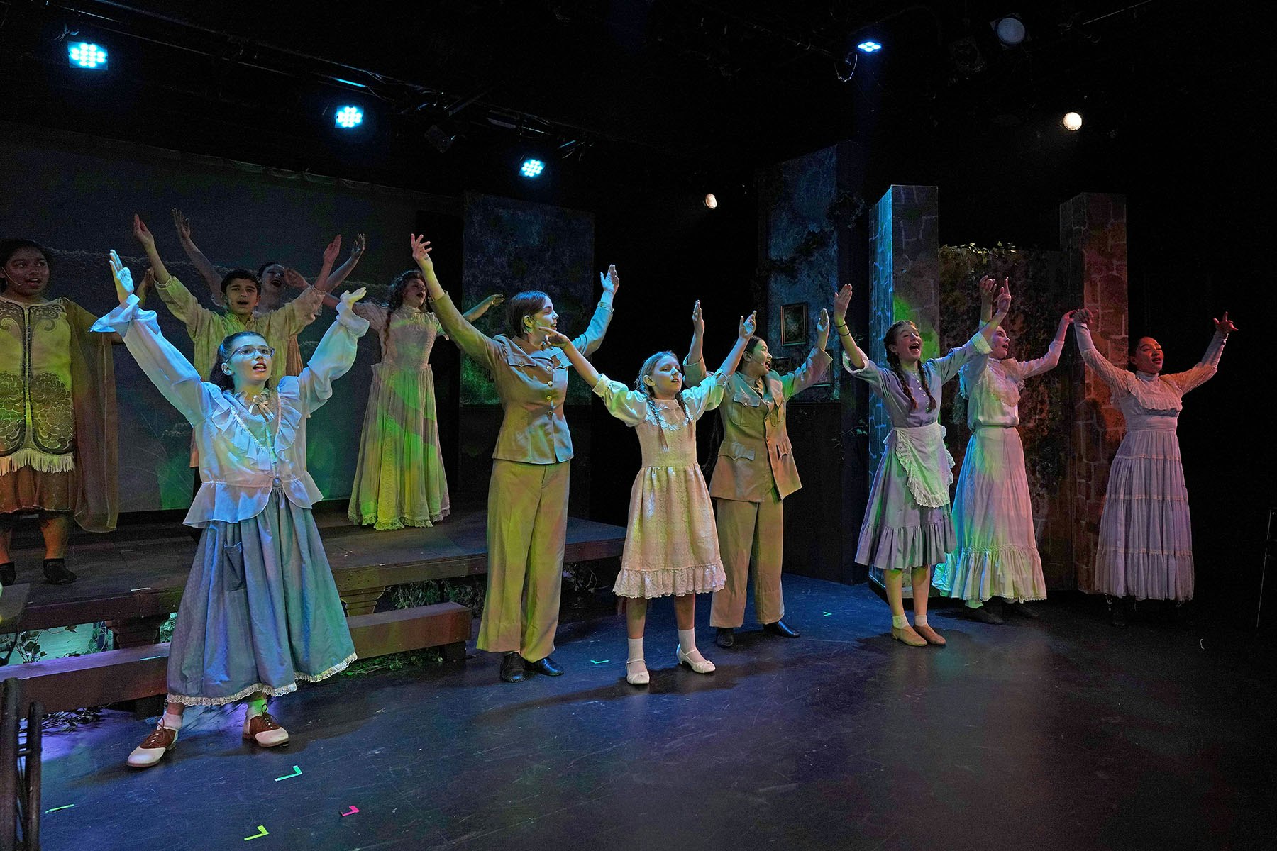 A group of performers on stage with vintage costumes, arms raised in a theatrical setting with dramatic lighting.