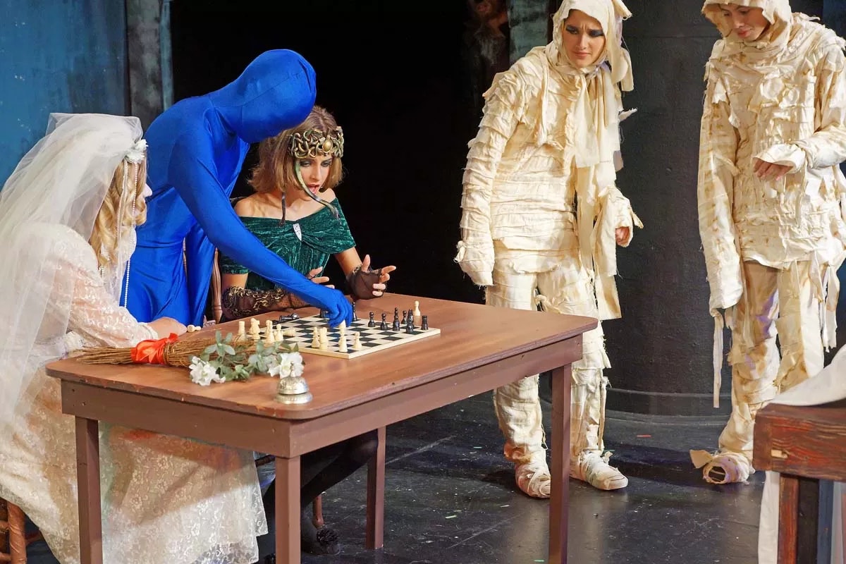 People in costumes, including a bride, mummies, and a blue figure, playing chess at a table.