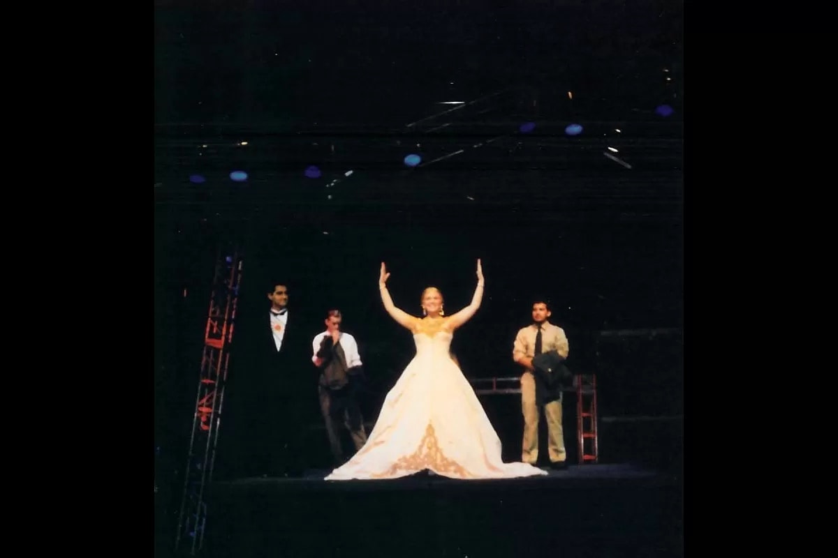 A woman in a gown stands on stage with arms raised, flanked by three men in formal attire.