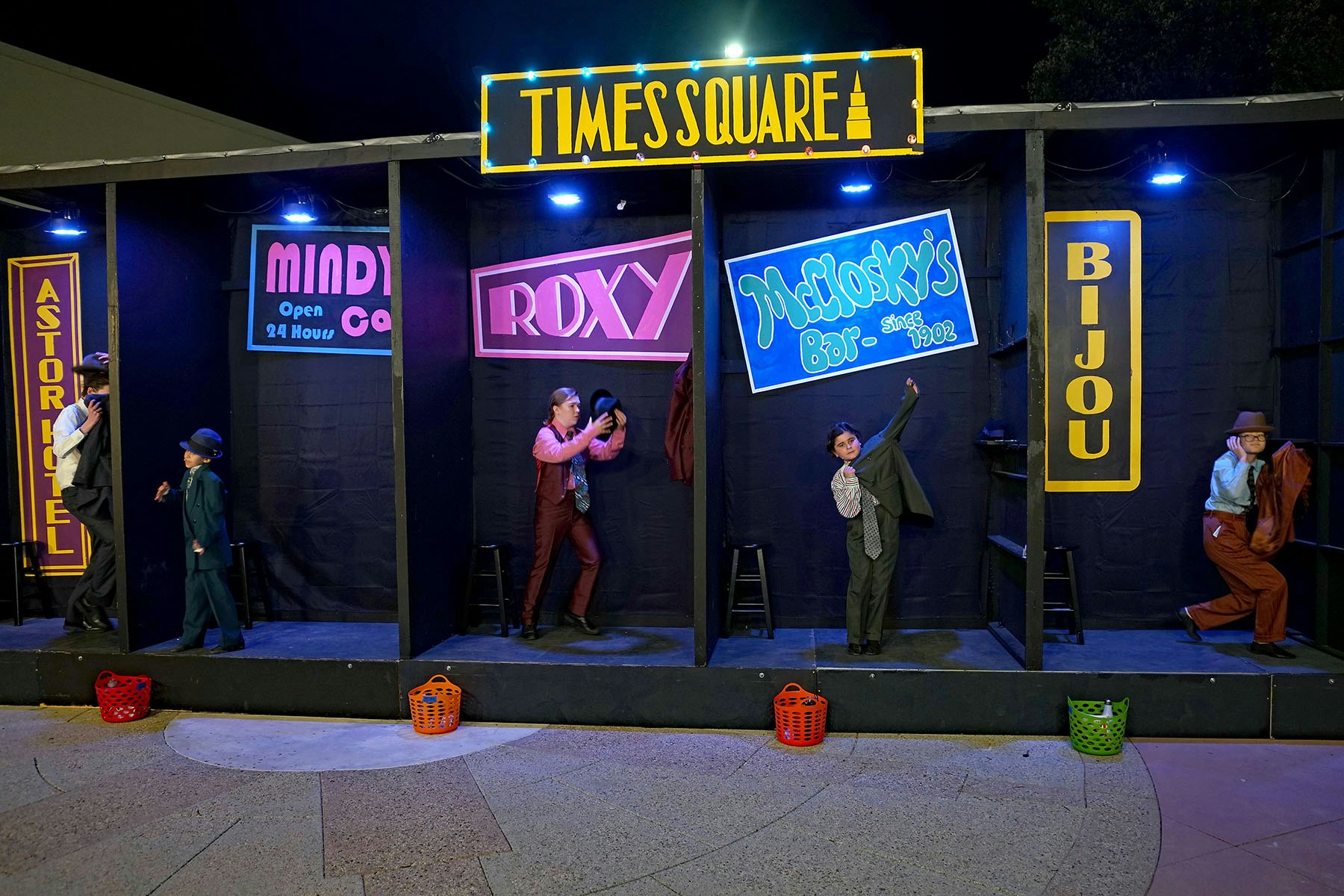 Children in suits on stage set resembling Times Square; colorful signs and baskets decorate the scene.