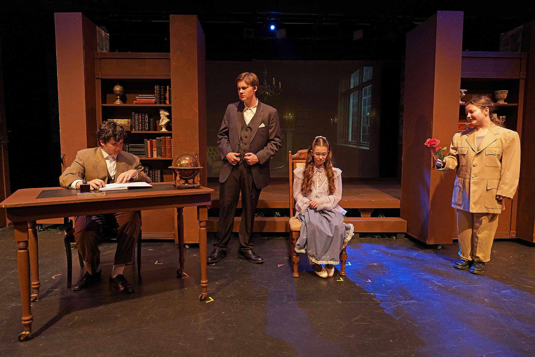 A theatrical scene with four actors on stage; one reads a book at a desk, another stands with a rose, others are standing and seated.
