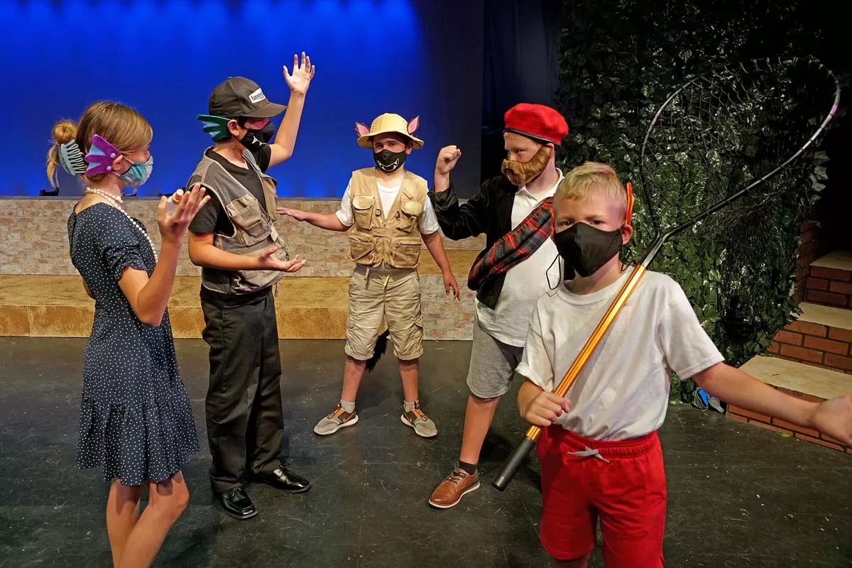 People in costumes, wearing masks, performing on stage with a blue backdrop and greenery.