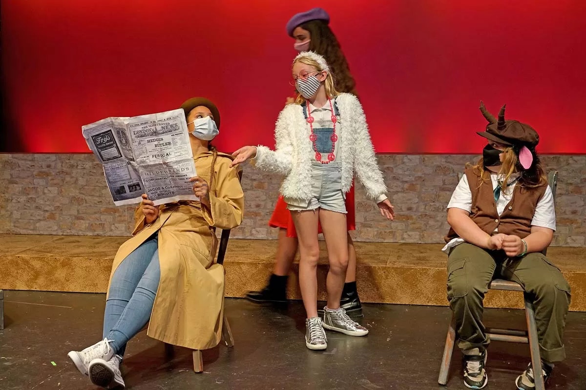 Four people in costumes and masks on stage; one reads a newspaper, another has a fluffy jacket, and a third wears horns.