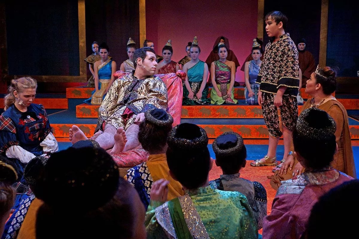 A group of people in colorful costumes on stage, depicting a theatrical performance with a character sitting on a throne.