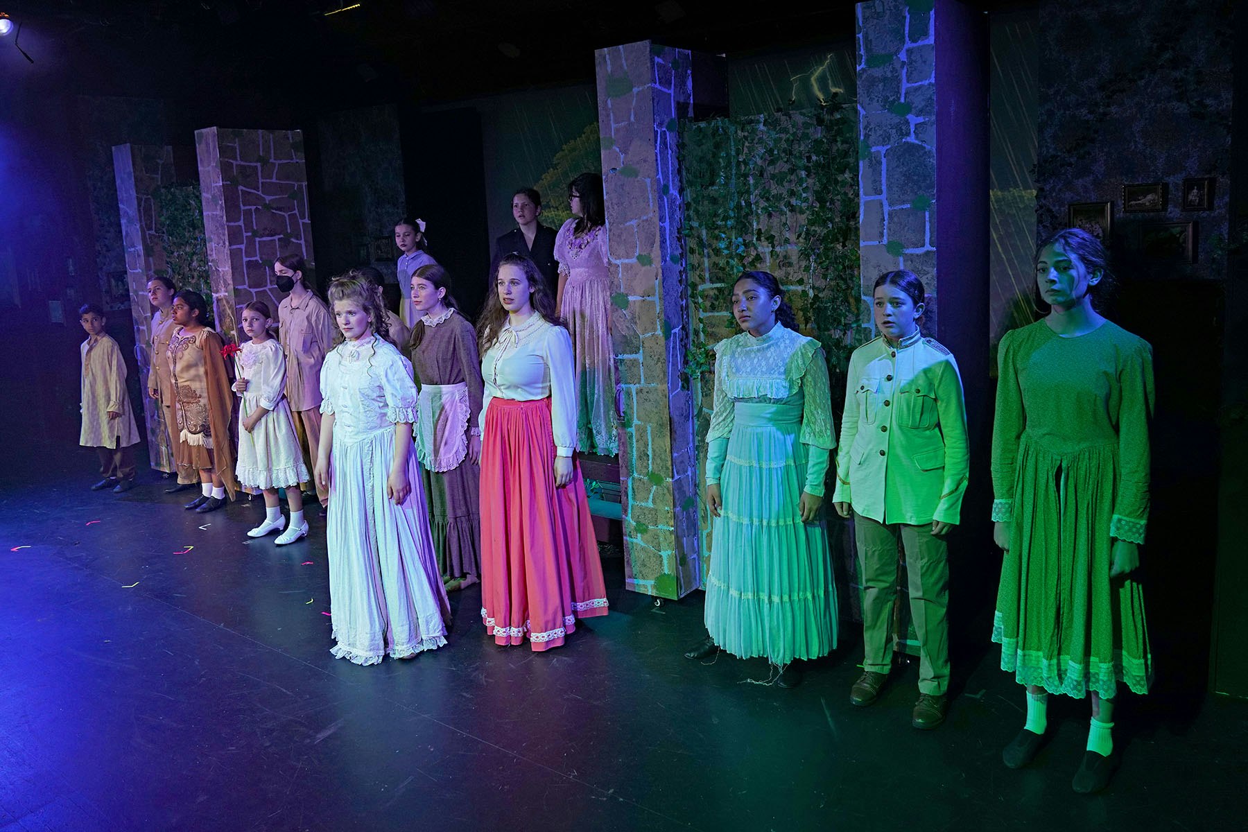A group of people in period costumes standing on stage with dramatic lighting.