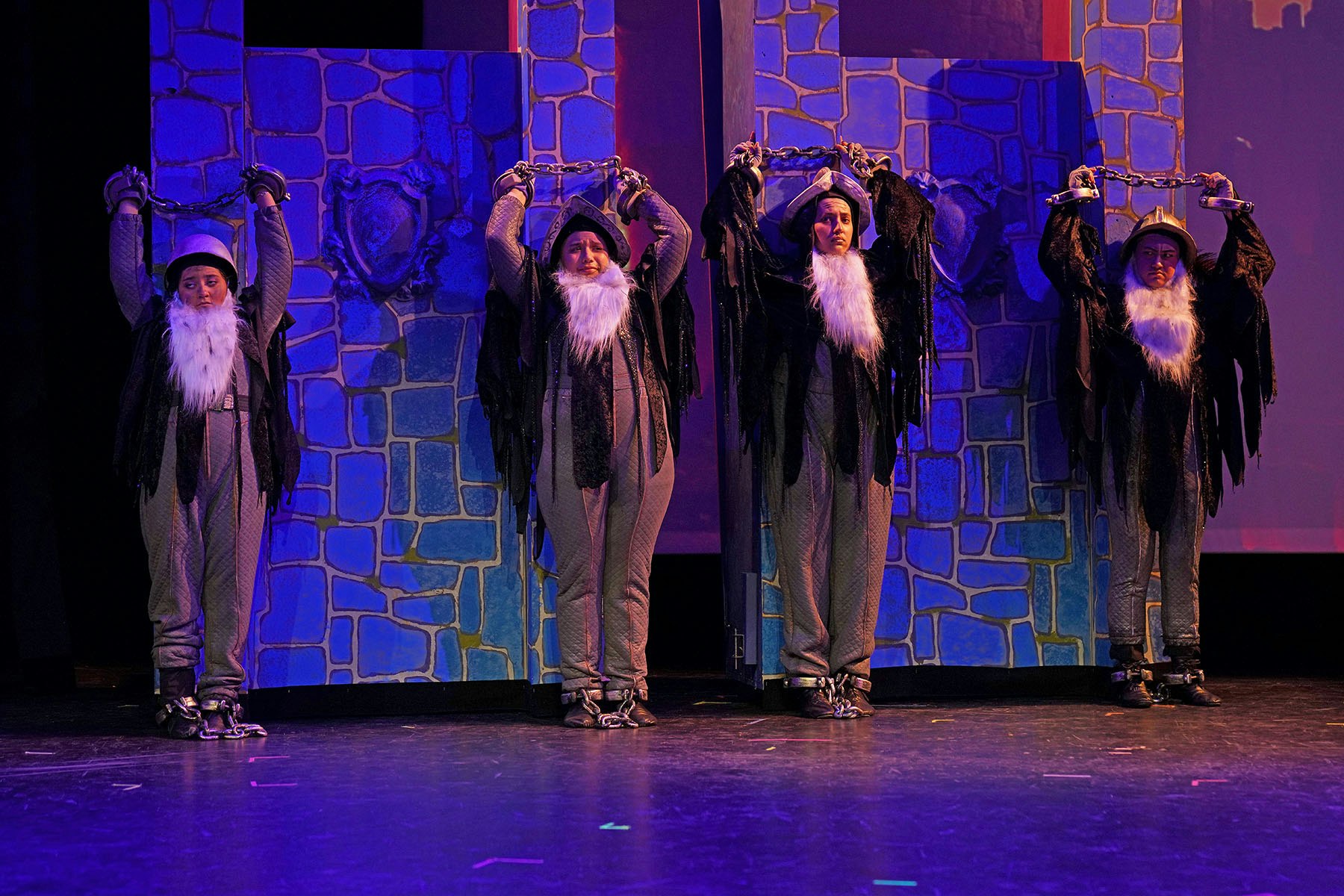 Four performers dressed as old men with beards, chained to a wall on stage.