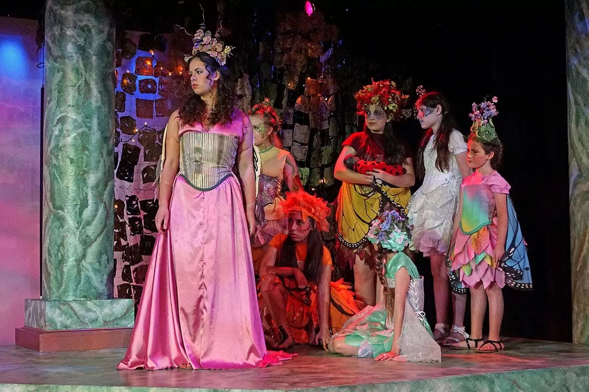 A group of people in colorful, fairy-like costumes on stage with decorative crowns and wings in a theatrical setting.