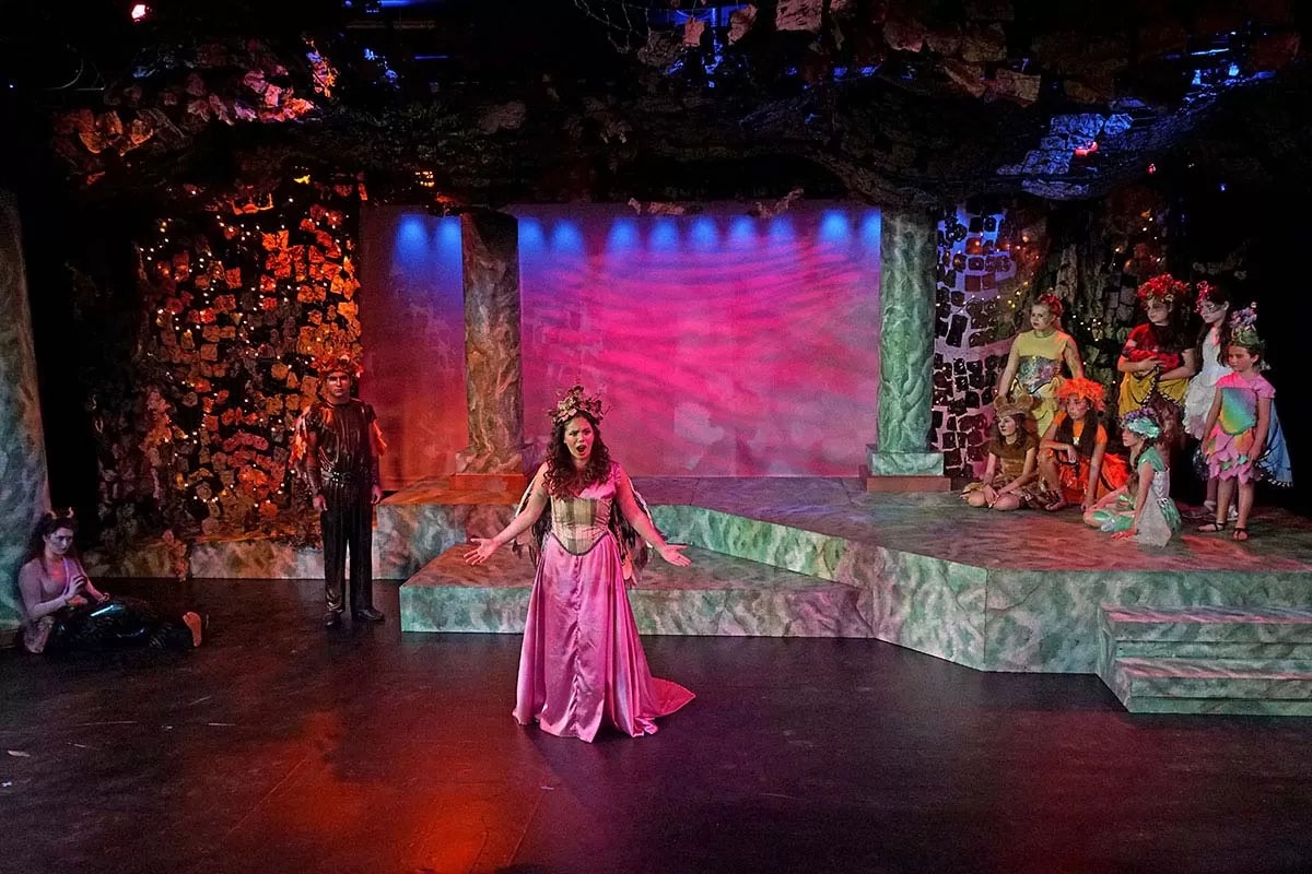 A theatrical stage with actors in colorful costumes performing a scene in a forest-like setting.