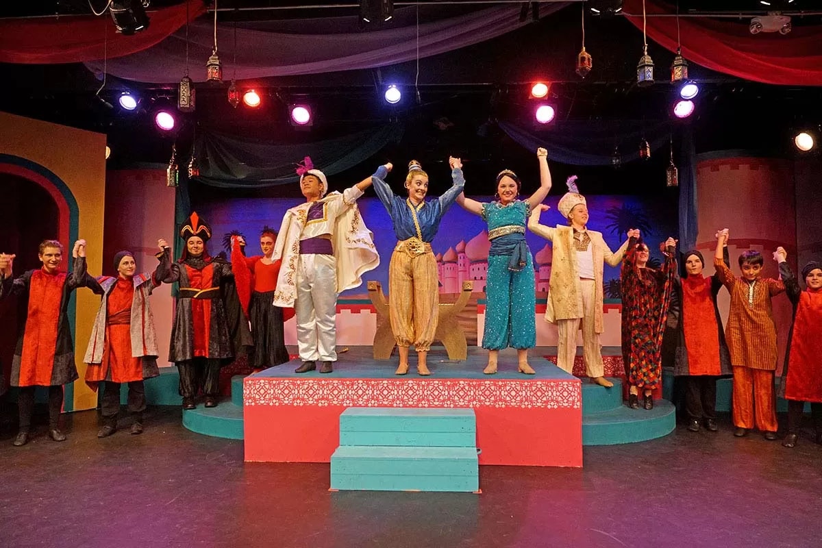 A group of actors in colorful costumes take a bow on a stage with a vibrant, Middle Eastern-inspired set design.
