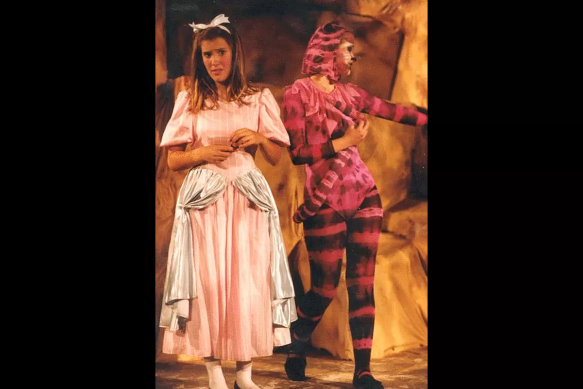 Two people in costumes: one in a pink dress with a bow, the other in a striped bodysuit. They appear to be in a theatrical play.
