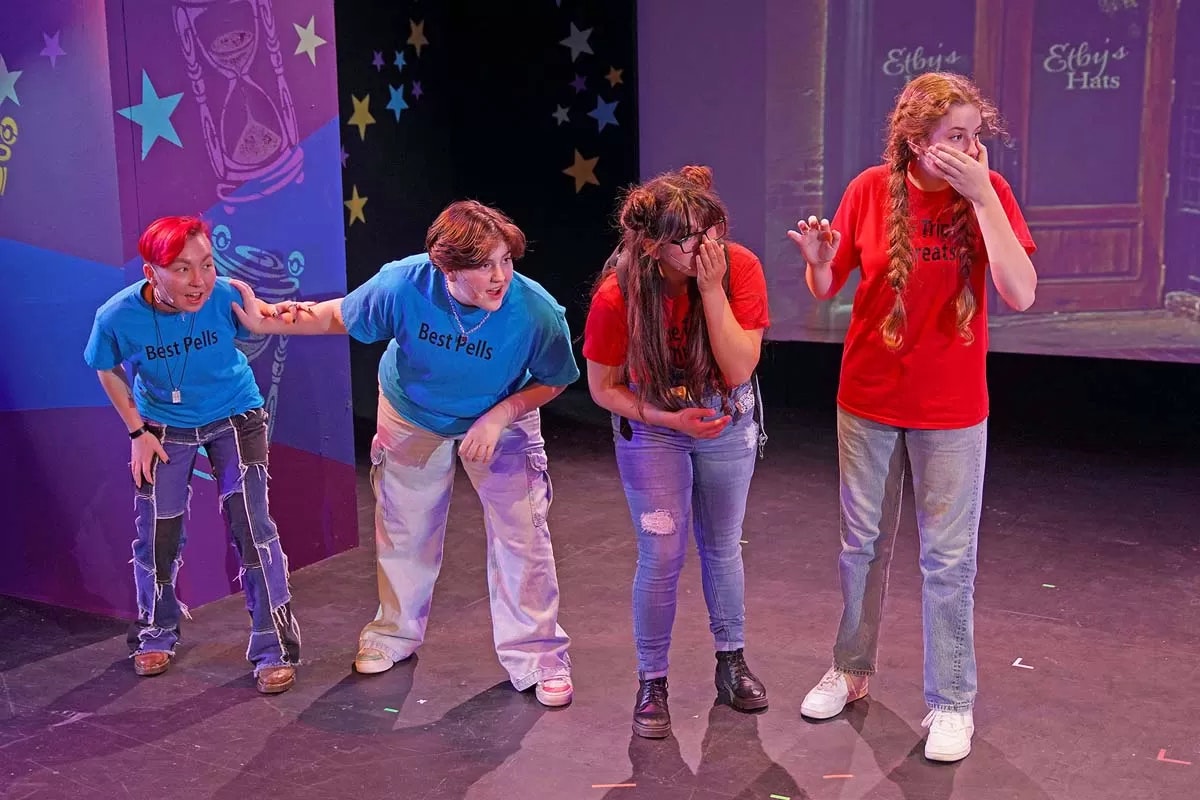 Four people on stage, two in blue "Best Pells" shirts, two in red shirts, appearing surprised or amused, with a colorful backdrop.