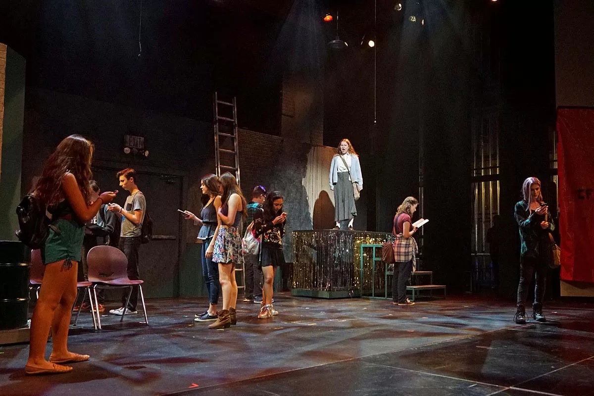 People on stage, mostly looking at phones, one person elevated speaking.