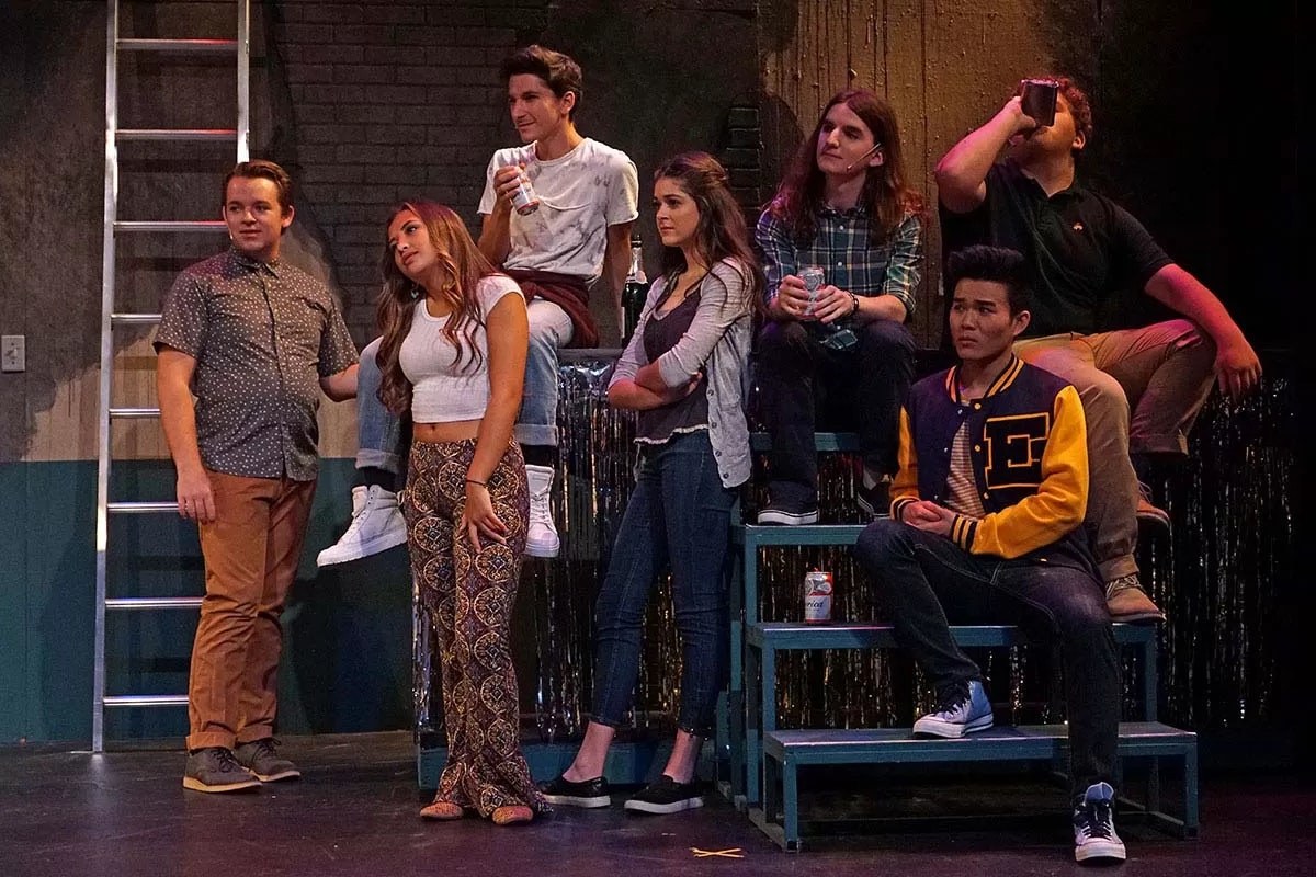 A group of seven people are casually gathered on stairs in a dimly lit room, some holding drinks and chatting.