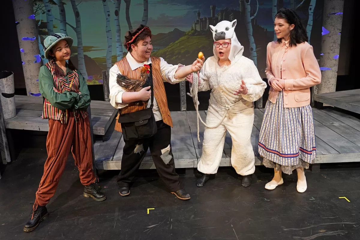 Four people in costume on a stage; one holds a chicken, another in an animal suit reacts to a golden egg.