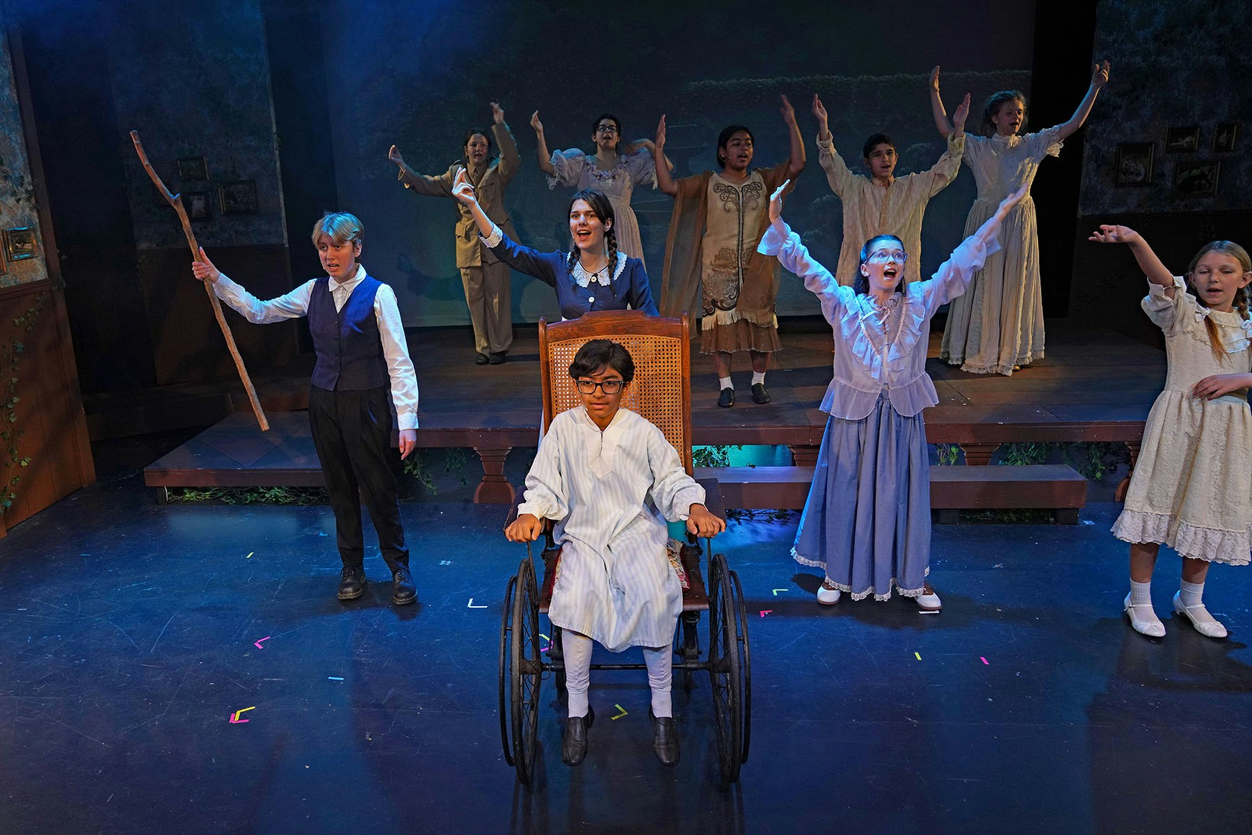 A group of actors performs on stage, with one in a wheelchair, others raising hands. The scene is theatrical and lively.