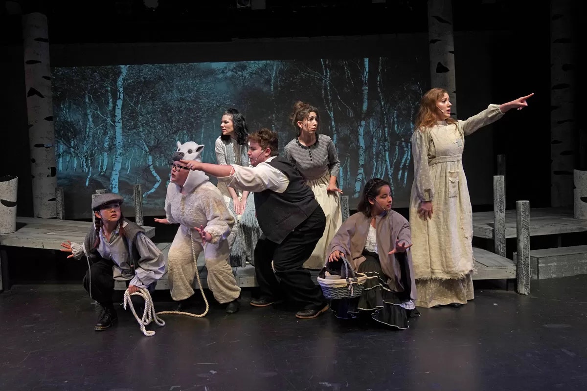 A group of actors in a theatrical forest scene, some pointing and posing dramatically, with one in animal costume holding a rope.