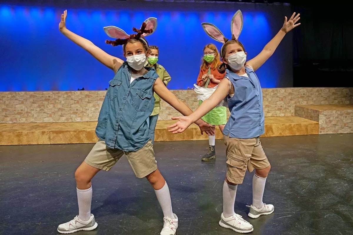 Kids on stage wearing masks, animal ears, and casual outfits, striking poses against a blue-lit background.