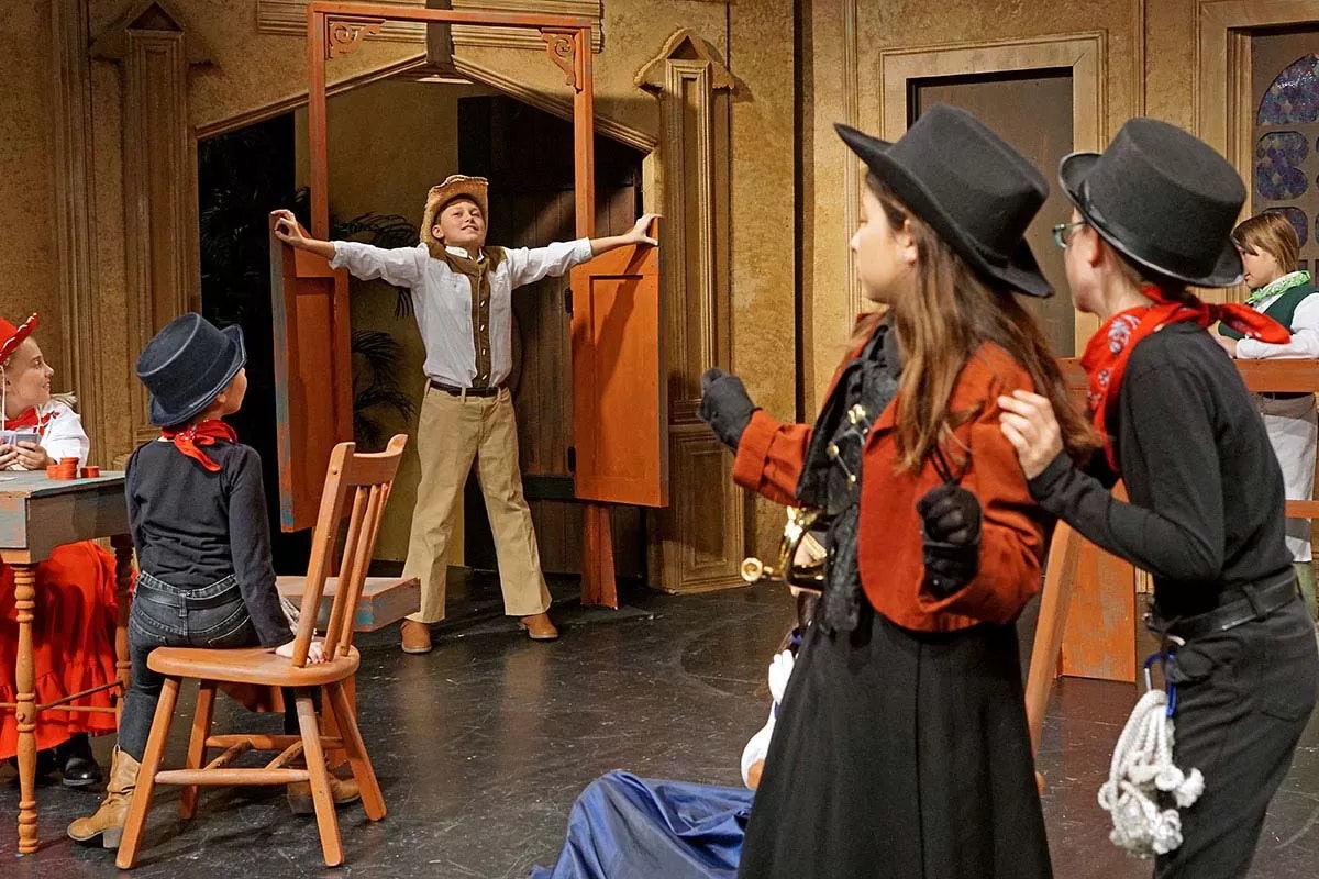 Children on stage in a Wild West-themed play; one in cowboy attire at saloon doors, others in hats watching.