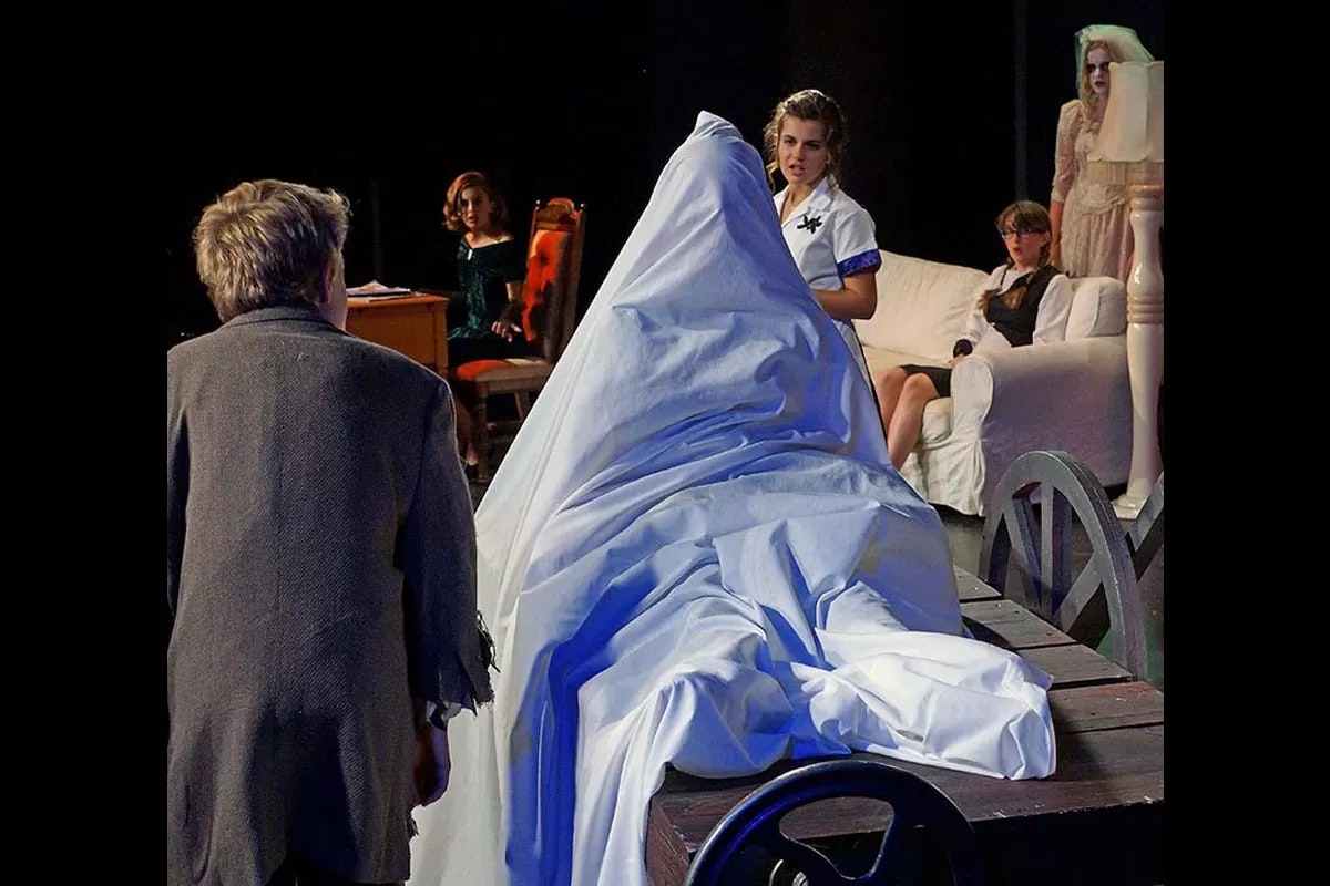 A scene from a play with actors: a person under a sheet, audience in costumes, including a ghostly figure in the back.
