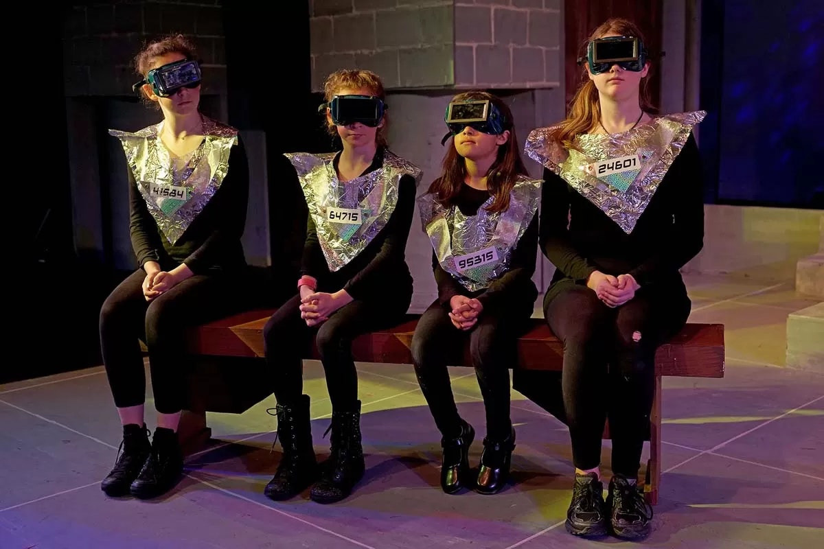 Four people in futuristic outfits with silver collar pieces and goggles, seated on a bench.