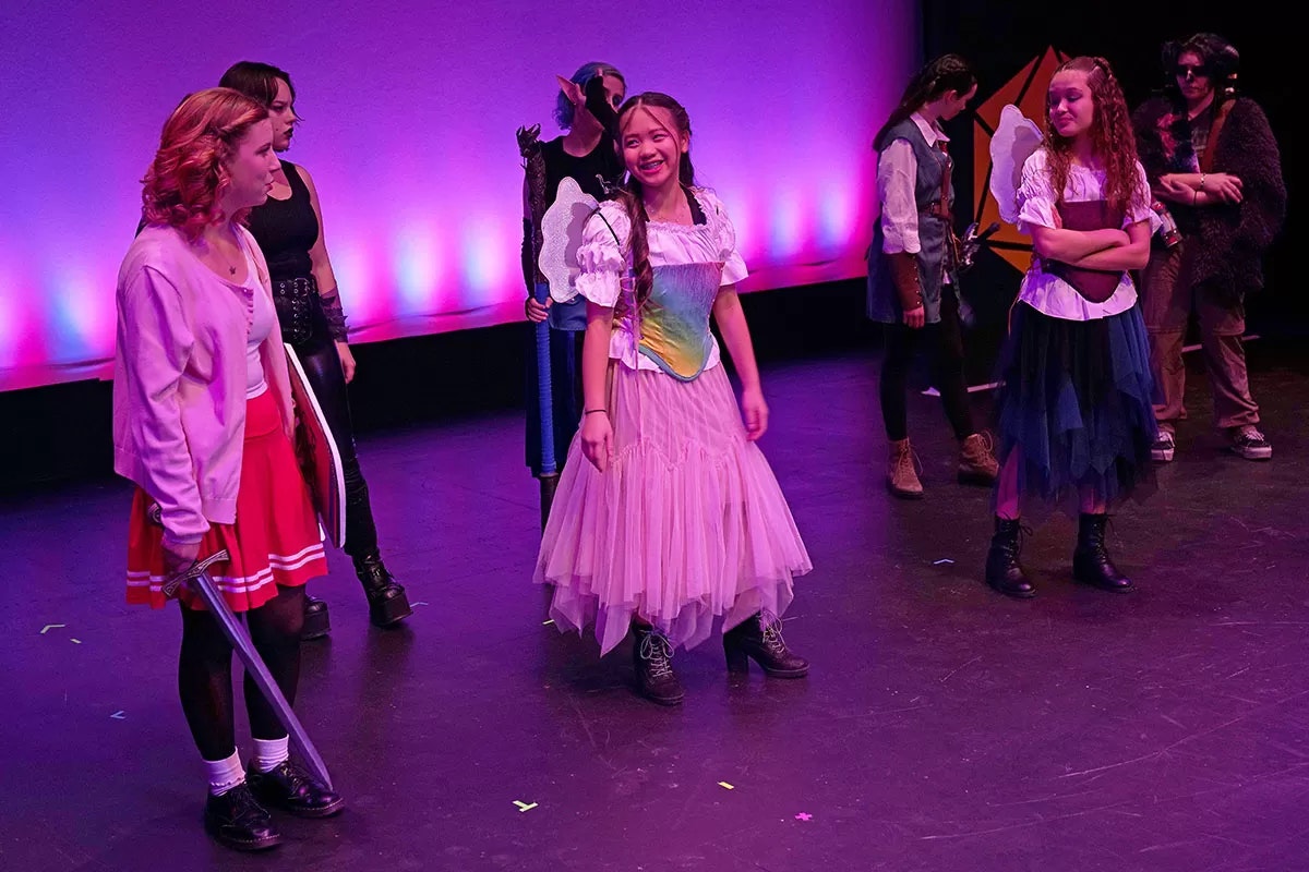 A group of people in colorful costumes is on stage, with a pink and purple-lit background.