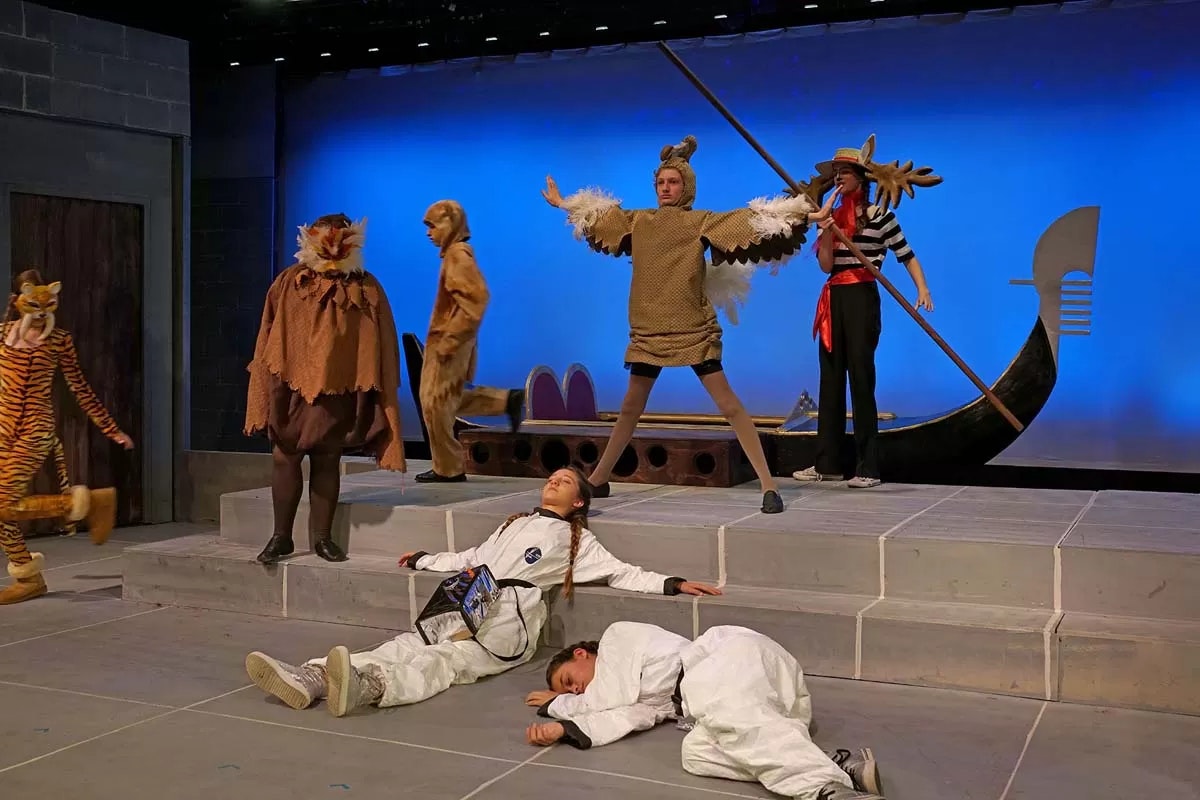 Performers in animal and costume outfits on a stage, with some lying and others standing, against a blue backdrop.