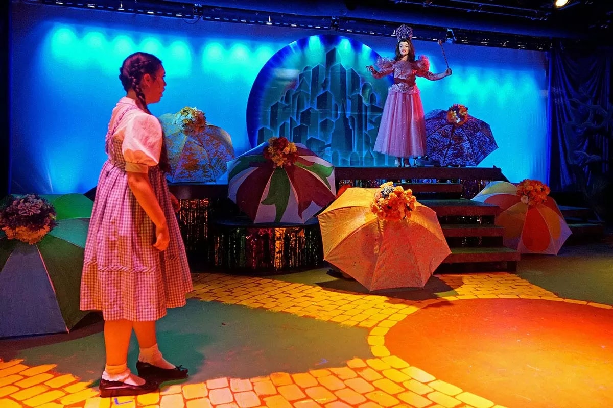 A colorful theater scene with two characters, one in a pink dress on a platform, surrounded by umbrellas and a yellow brick road.