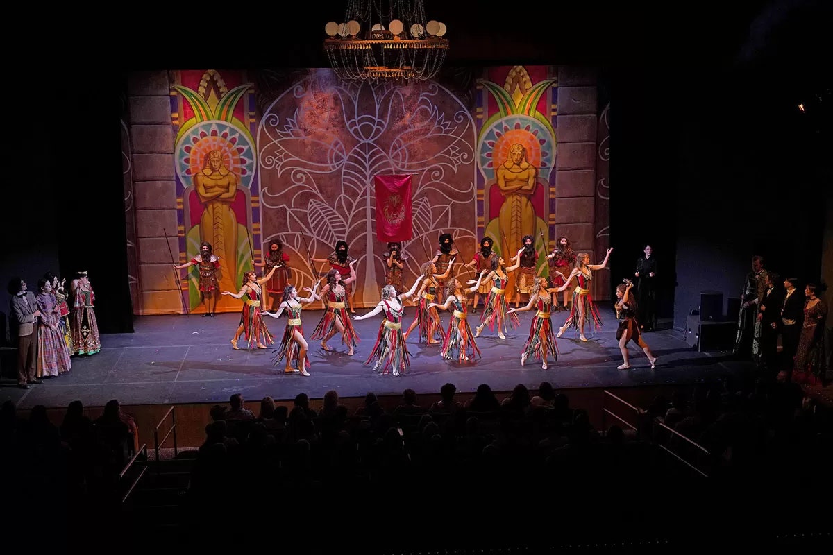 A vibrant stage performance with dancers in colorful costumes and an elaborate backdrop featuring artistic designs and statues.