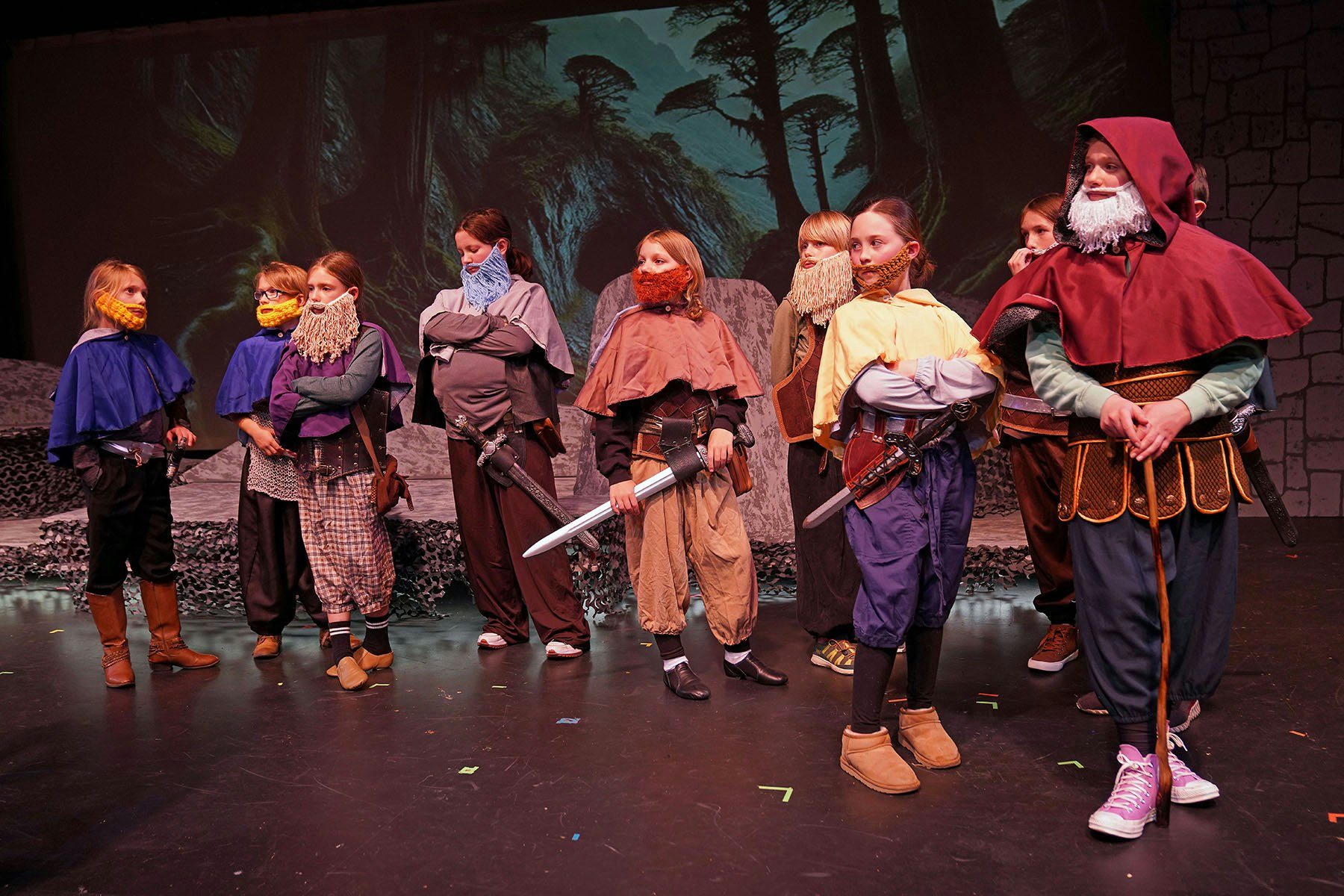 A group of children dressed as fantasy characters with beards and capes, holding swords on a stage with a forest backdrop.